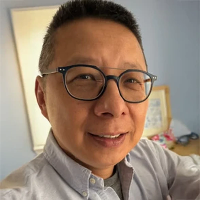 A close-up headshot of a smiling East Asian person with short dark hair. They are wearing dark-rimmed glasses and a light blue button-down shirt.