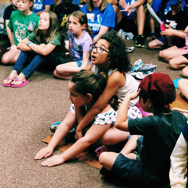 A diverse group of young children sit on a carpeted floor, looking up with engaged and wondrous expressions at something happening out of frame.