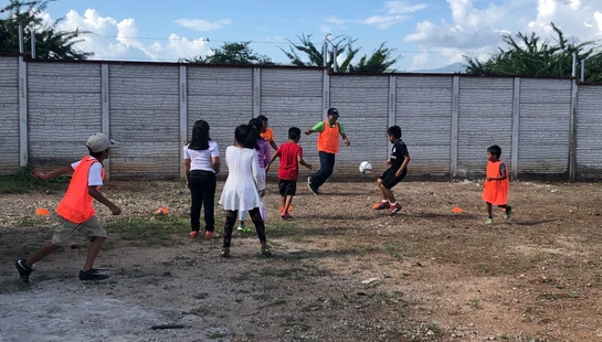 A group of children and an adult play soccer on a dirt field in front of a large concrete wall. Some players wear bright orange vests.