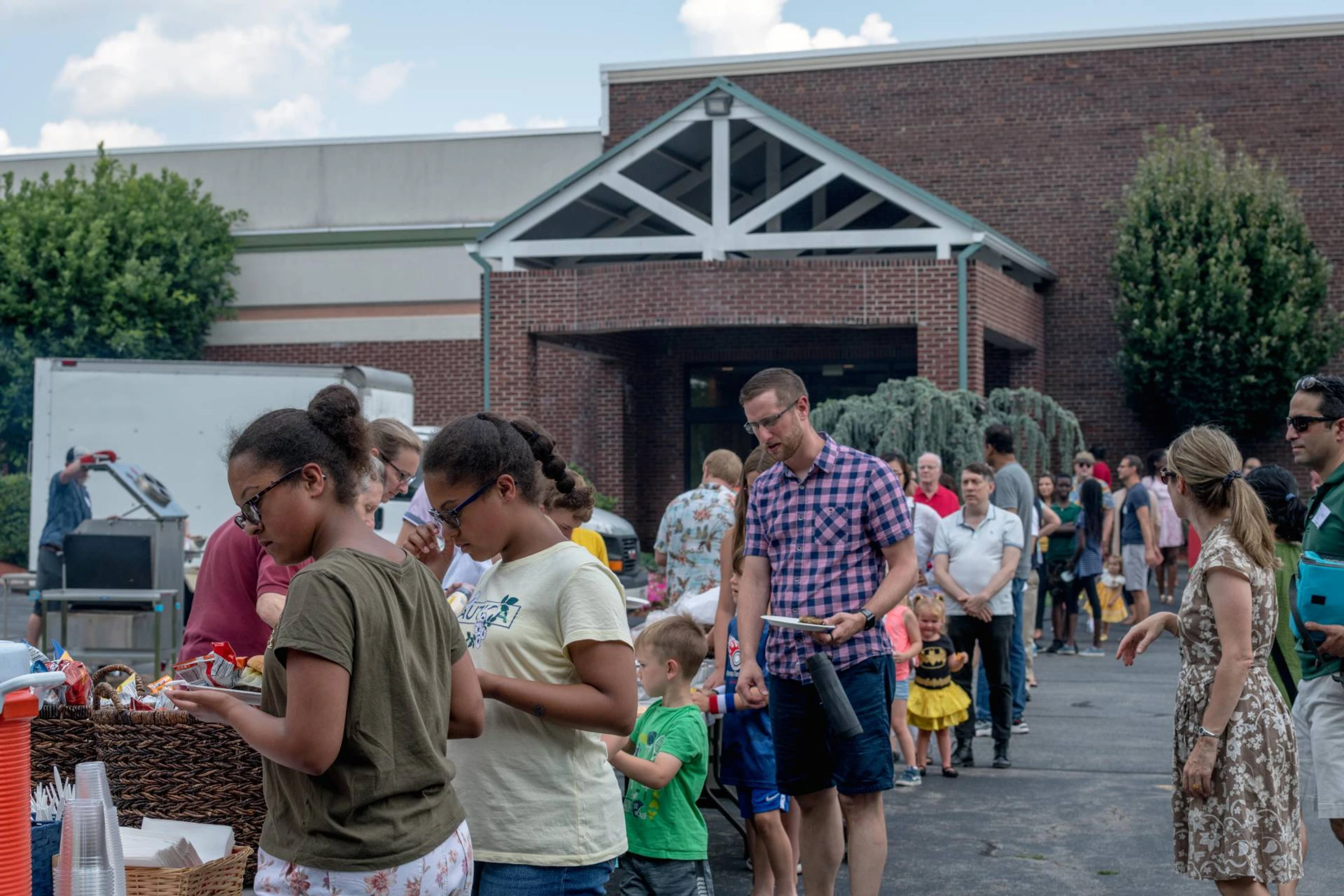 A diverse group of adults and children stand in line for food at an outdoor community cookout in a parking lot in front of a brick building.