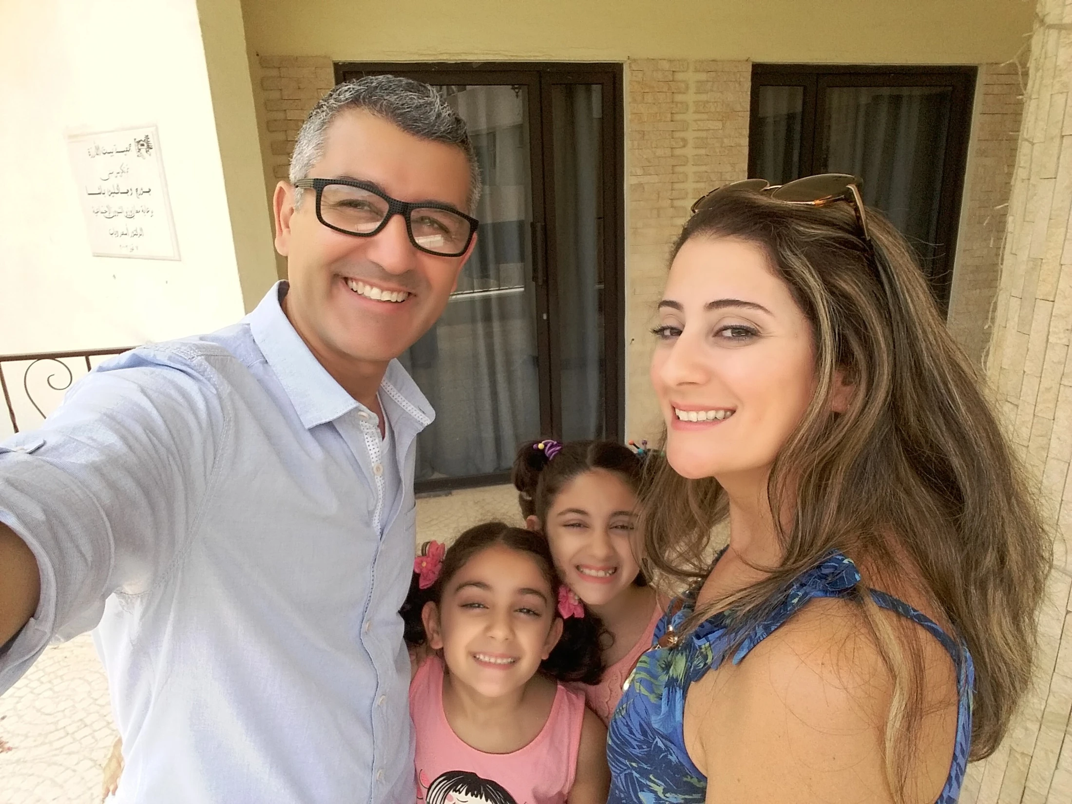 A smiling family of four takes a selfie outdoors. A man with glasses and a woman pose with their two young daughters who are smiling between them.