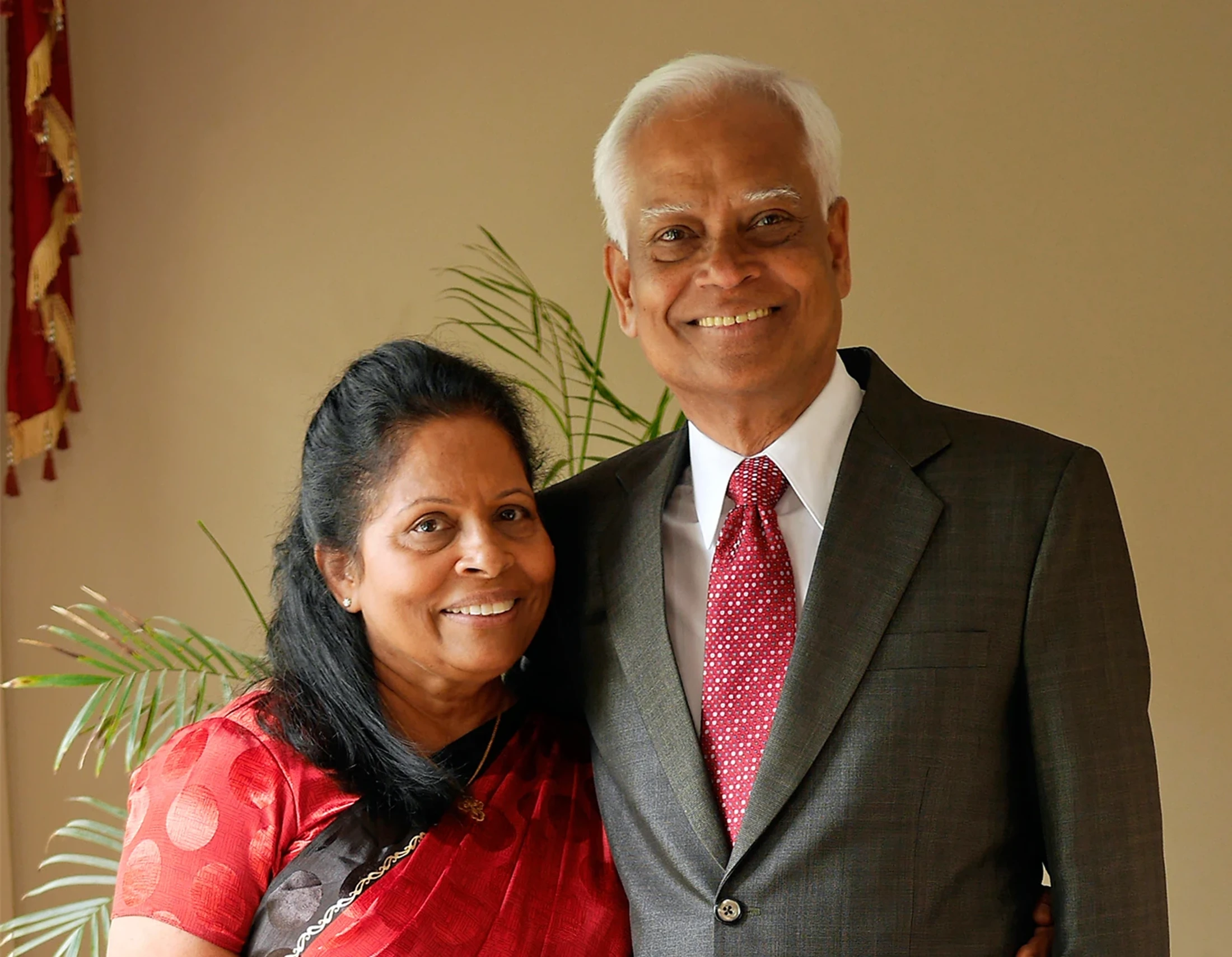 An older couple of South Asian descent smiling at the camera. The woman wears a red sari, and the man wears a dark suit with a red patterned tie.