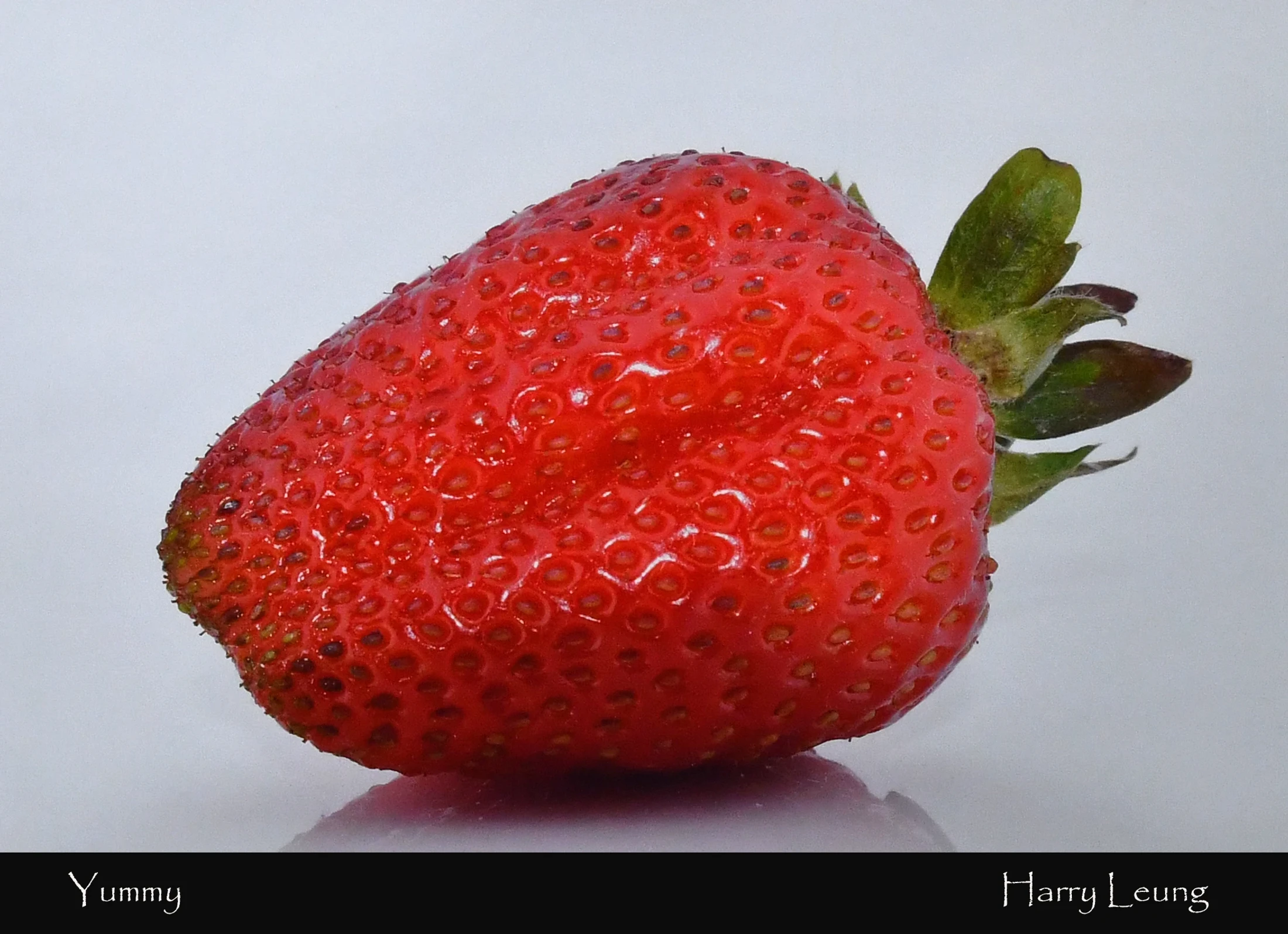 A detailed close-up shot of a single, shiny red strawberry with its green calyx intact, resting on a reflective white surface against a plain background.