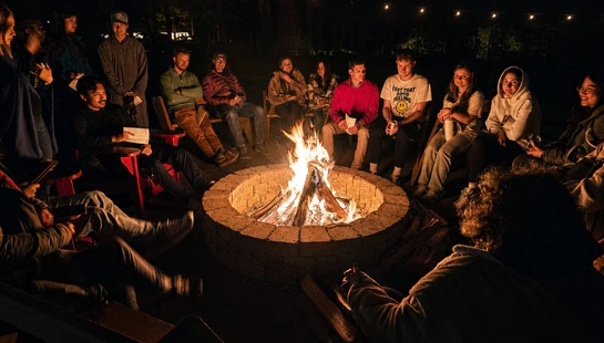 A diverse group of young adults sits in a circle around a blazing bonfire in a brick fire pit at night, illuminated by the flames and string lights above.
