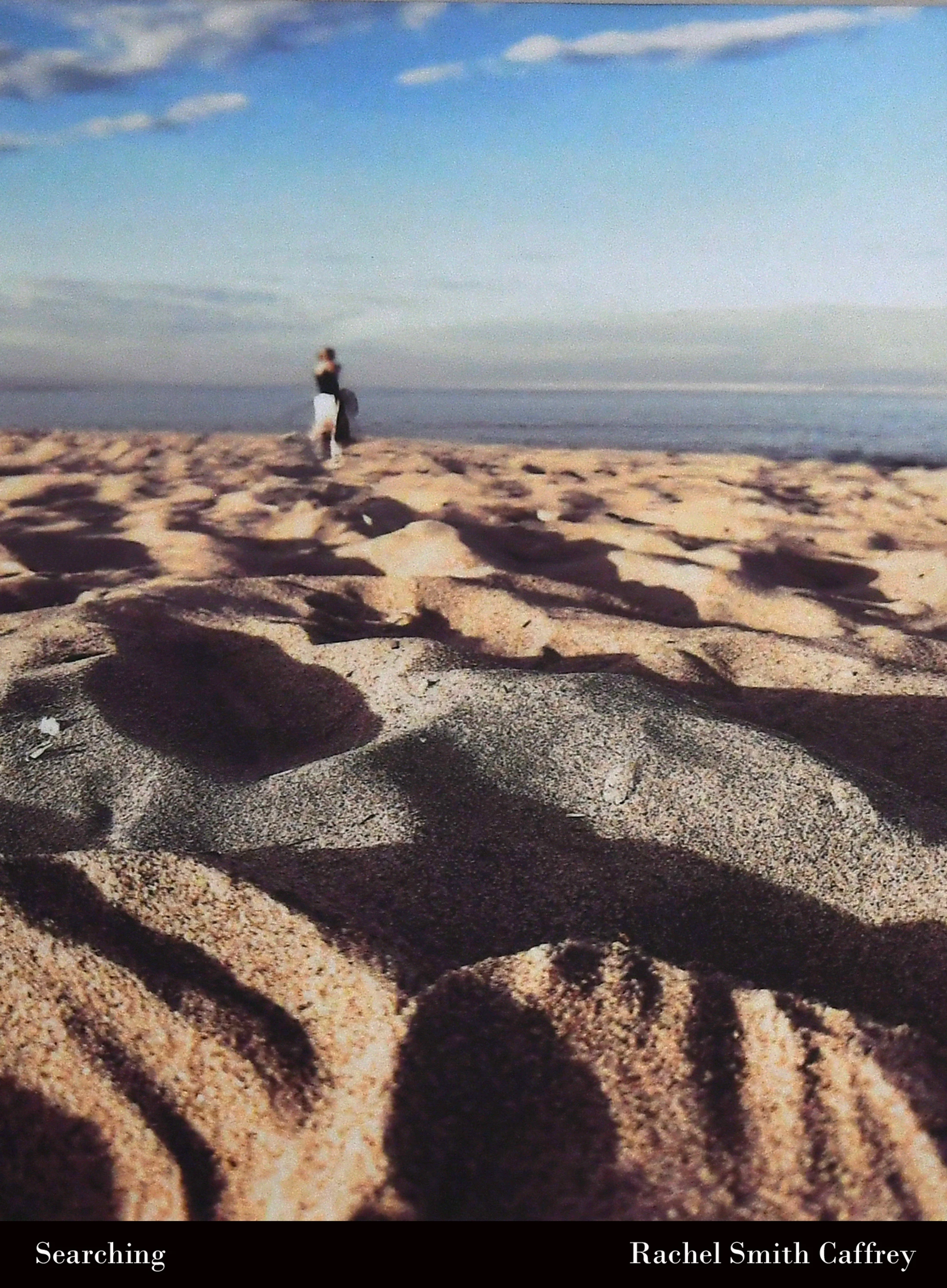 A low-angle, close-up shot of sand on a beach, with a lone, blurry figure standing by the ocean in the distance under a blue sky.