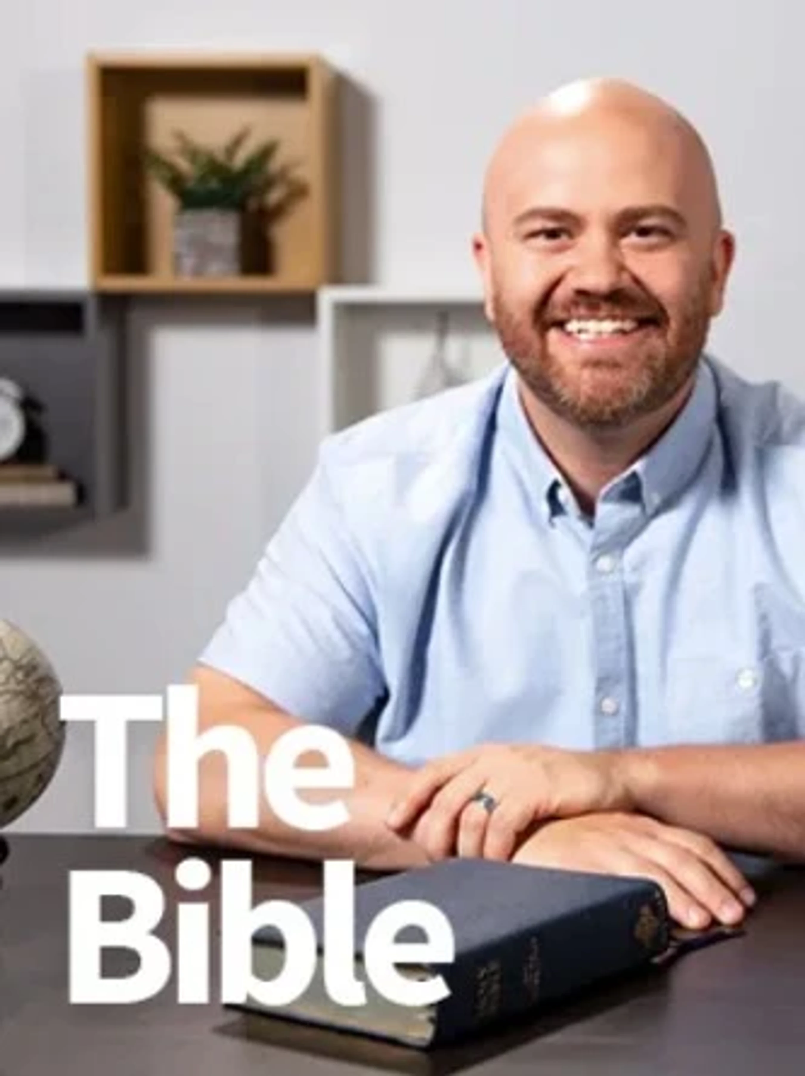A smiling, bald man with a beard sits at a desk with his hands near a blue Bible. The words 'The Bible' are written in large white letters in the foreground.