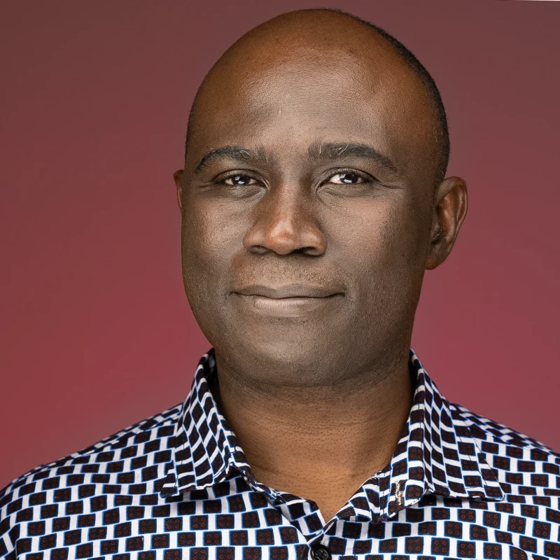 A studio headshot of a bald Black man with a gentle smile, wearing a black and white patterned collared shirt against a solid maroon background.