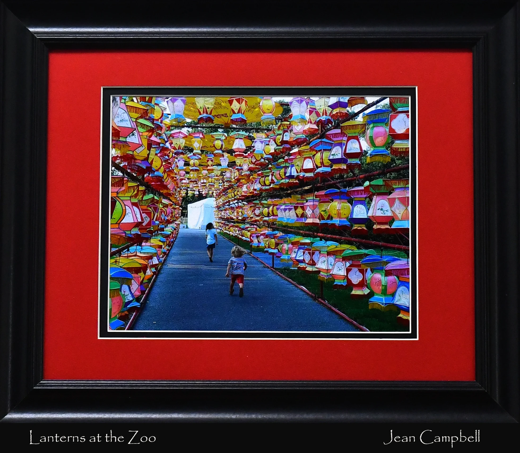 A framed photograph of two children running down a path lined on both sides with hundreds of colorful, decorative paper lanterns creating a tunnel effect.
