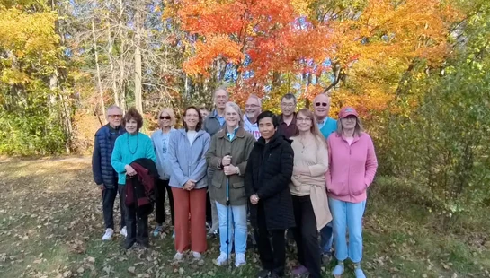 A group of twelve adults stand together for a photo in a park. Behind them, trees show vibrant orange, yellow, and red autumn leaves.