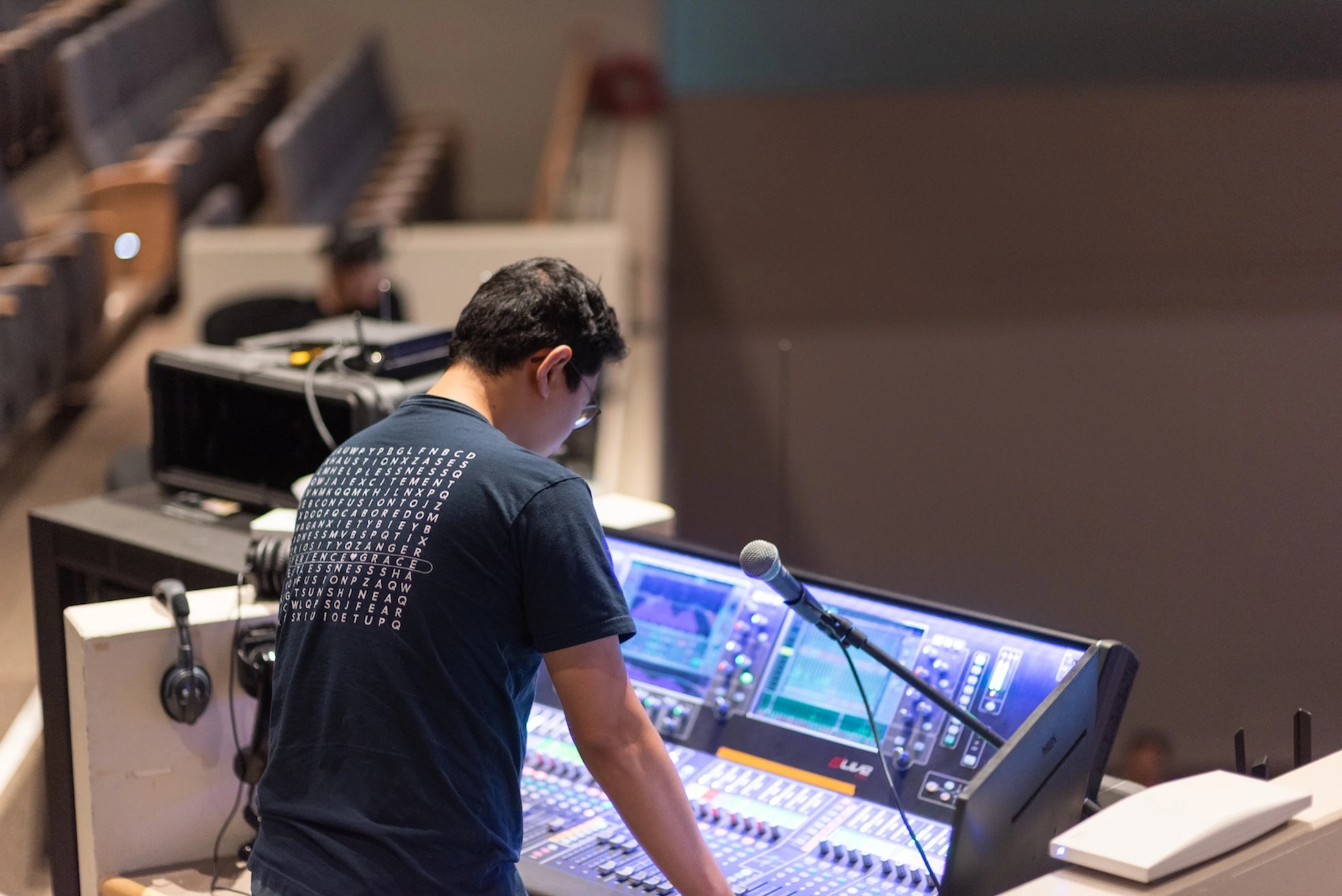 A man from behind, wearing a blue t-shirt, stands over a large, illuminated audio mixing board in a theater or auditorium, adjusting the controls.