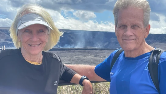 An older man and woman in athletic wear smile for the camera, posing in front of a vast volcanic crater with steam rising from it under a cloudy sky.