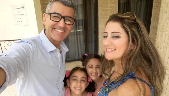 A smiling family of four takes a selfie outdoors. A man with glasses and a woman pose with their two young daughters who are smiling between them.