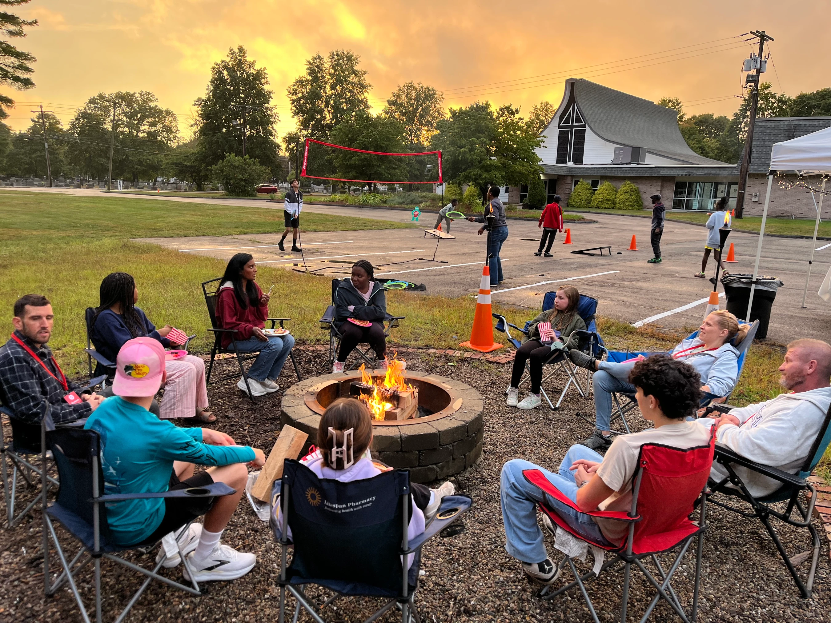 A group of adults and teens sit in chairs around a fire pit at dusk. In the background, others play volleyball and lawn games in a parking lot.
