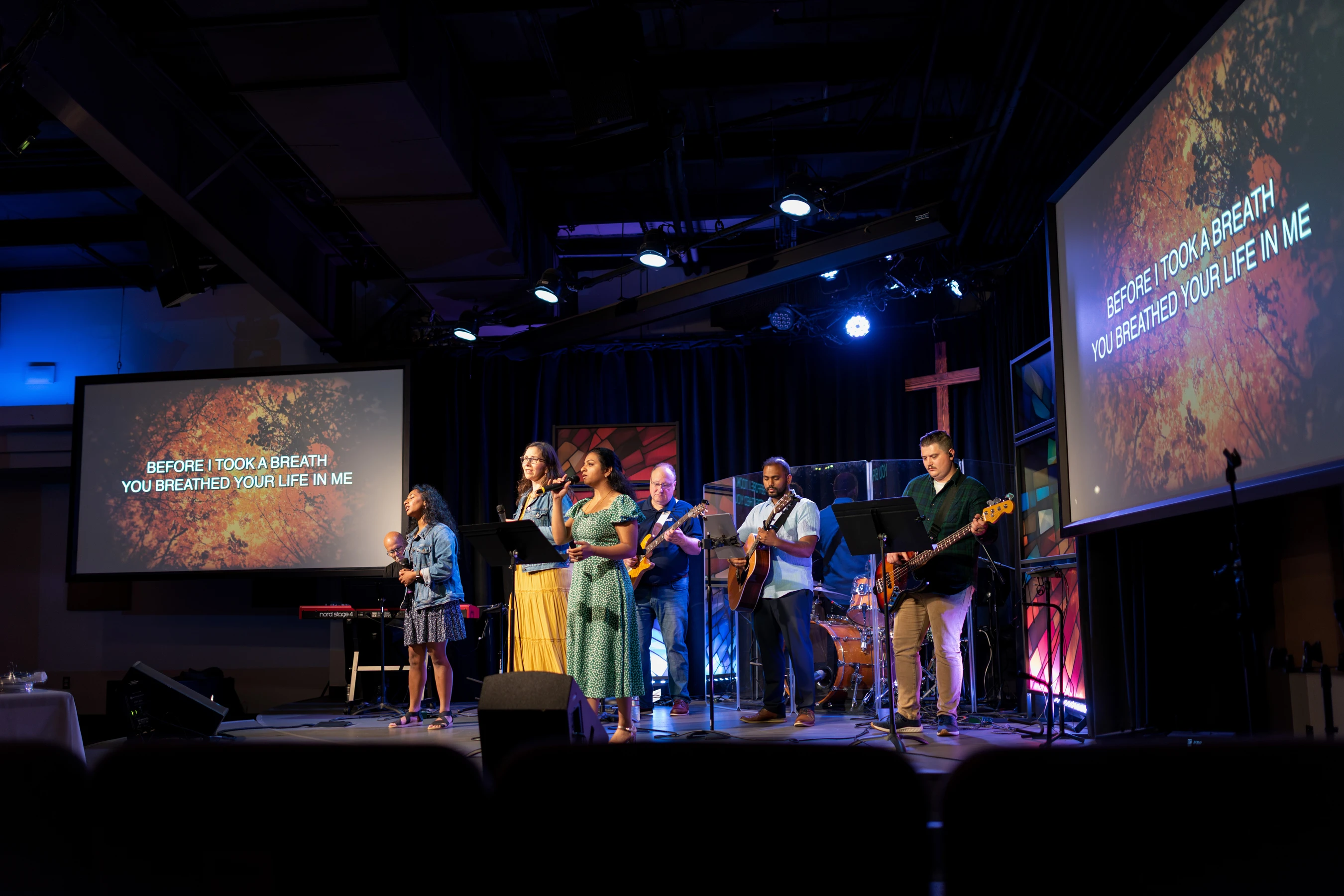 A diverse church worship band sings and plays instruments on a dimly lit stage, with lyrics displayed on large screens on either side.