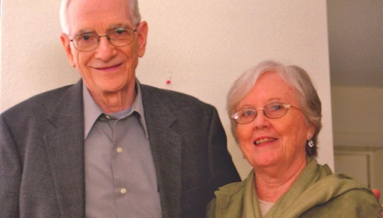 A waist-up portrait of an elderly man and woman standing side-by-side. He wears a grey blazer and she wears a green jacket. Both have white hair and glasses.