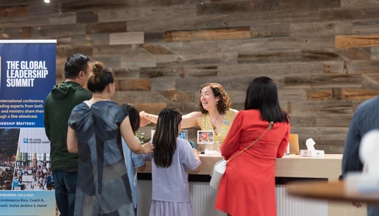 A smiling woman at a registration desk warmly greets a family, shaking a young girl's hand, next to a banner for The Global Leadership Summit.
