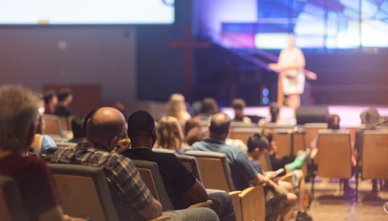 A view from behind of people sitting in auditorium seats, listening to a speaker on a brightly lit stage during a service or presentation.