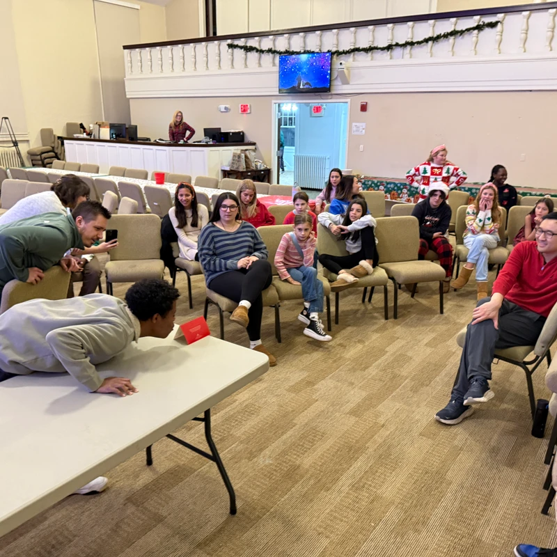 A diverse group of people watches a young man play a game, blowing on a red card across a table during a holiday gathering in a large hall.