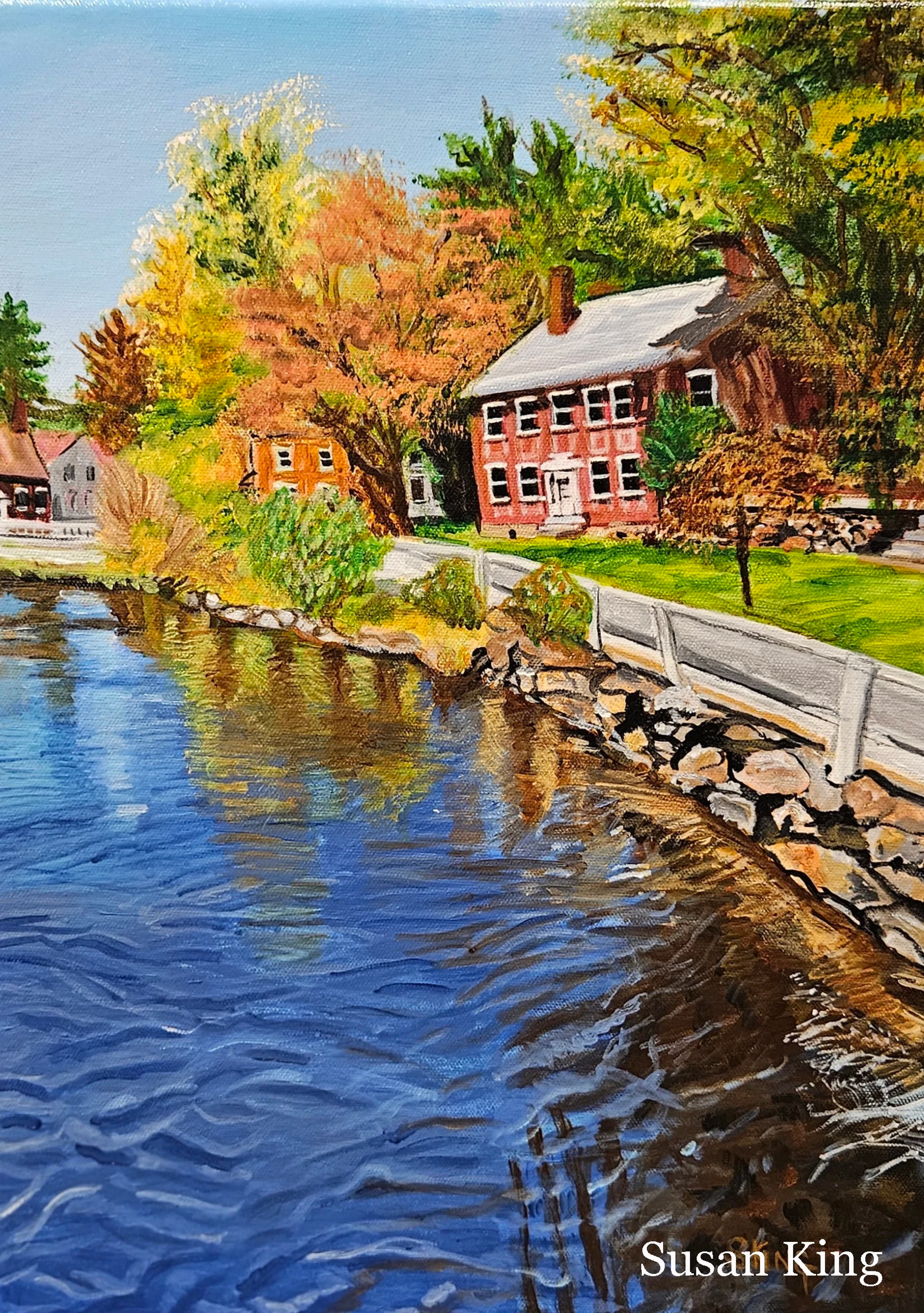 A vibrant painting of a riverside village in autumn. A large red colonial house sits on the bank next to blue water reflecting the colorful fall foliage.