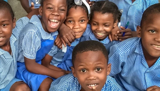 A group of young Black schoolchildren in blue and white checkered uniforms smile and laugh together while posing for a photo.