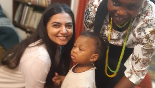 A smiling woman with long dark hair poses with a young child, while another woman in a floral blouse smiles down at them from behind.