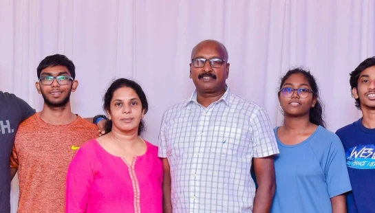 A family portrait of six people, two parents and four young adult children, standing together and smiling in front of a white curtain backdrop.