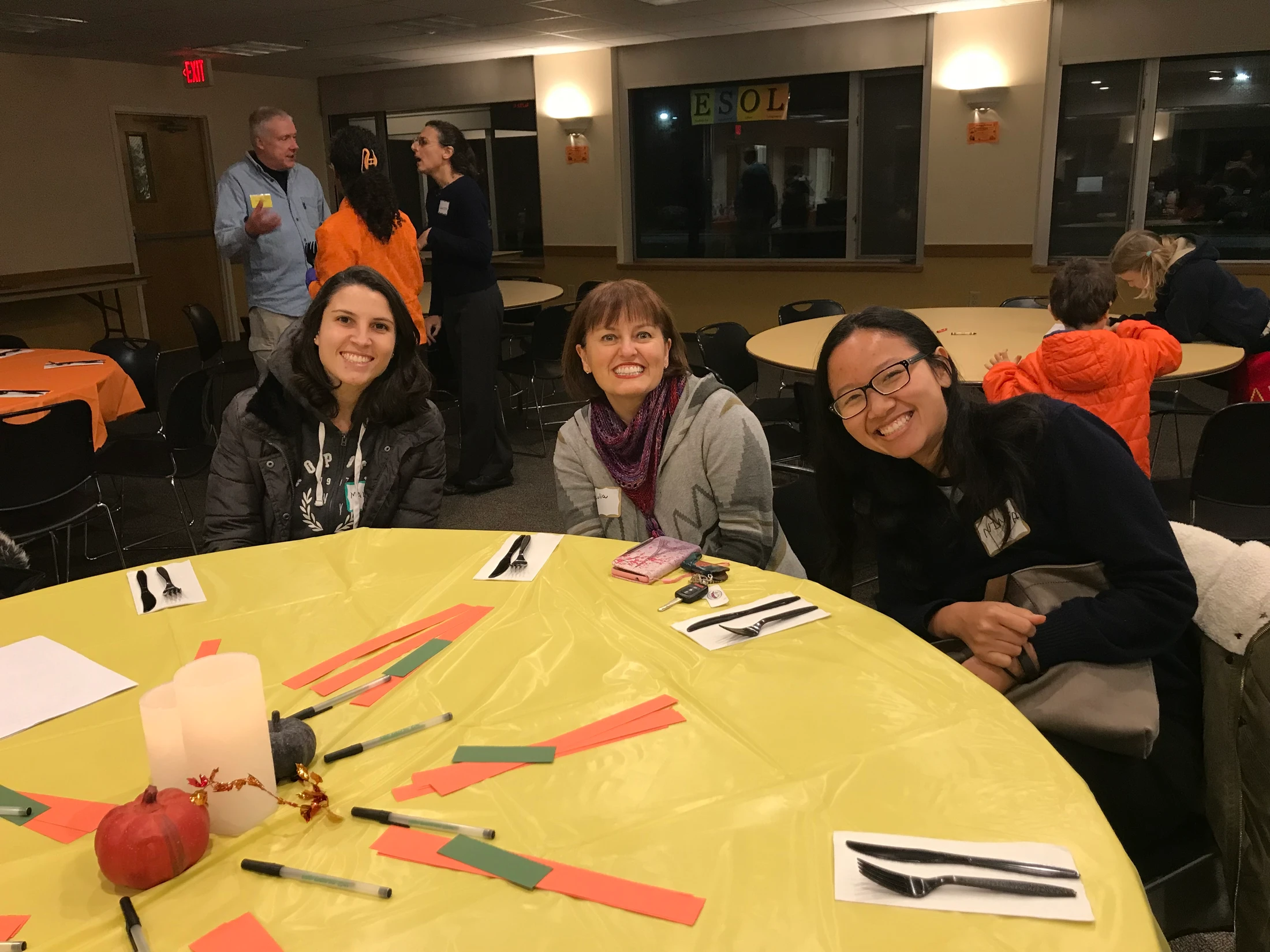 Three women smile while sitting at a round table with a yellow tablecloth at an indoor community event. Other people are mingling in the background.
