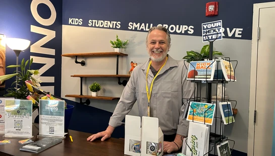 A smiling man with a gray beard stands behind a welcome desk at a church. The wall behind him says 'Connect' and lists options like 'Kids' and 'Small Groups'.