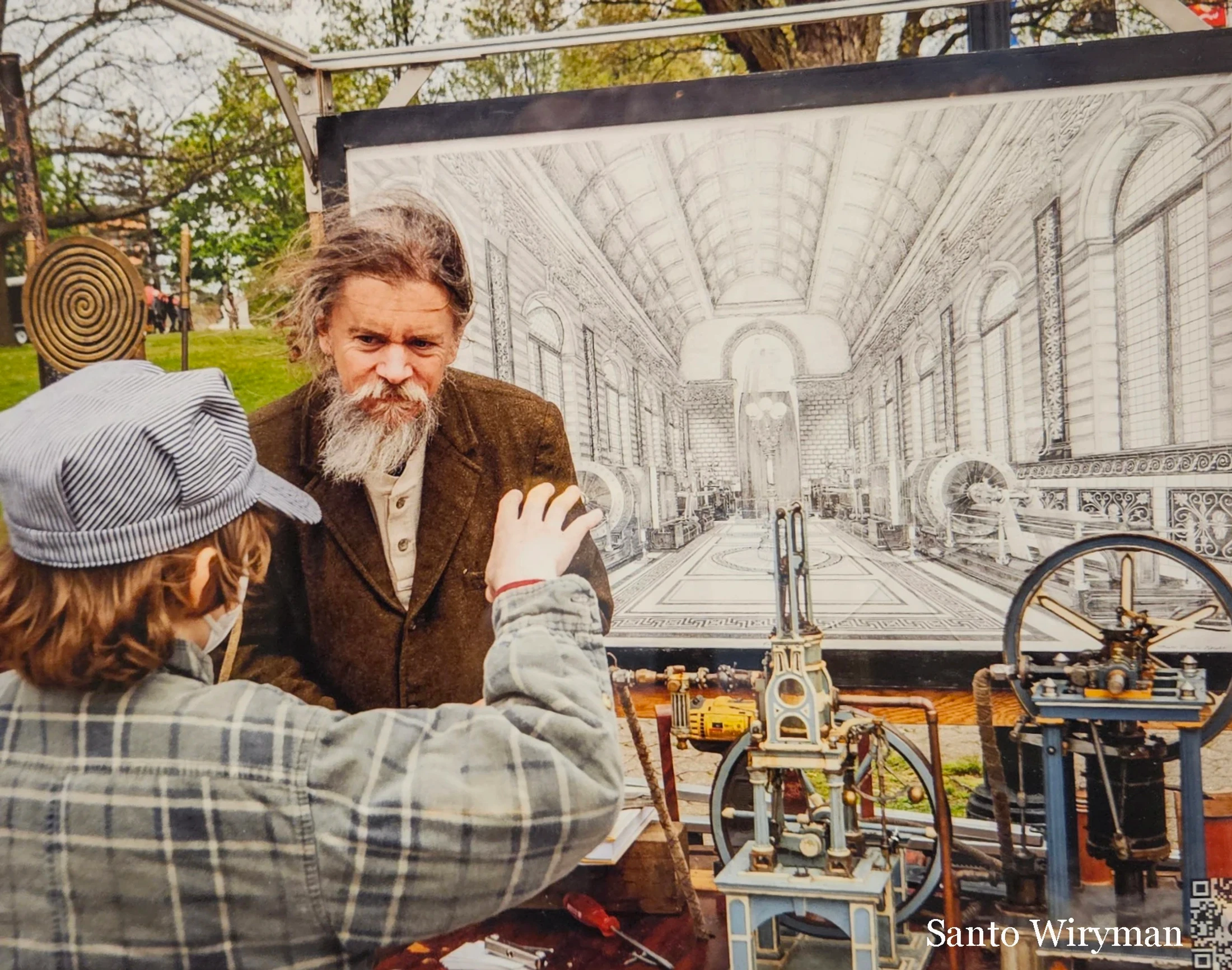An older man with a long grey beard explains his intricate mechanical models to a young person in a striped cap at an outdoor fair.