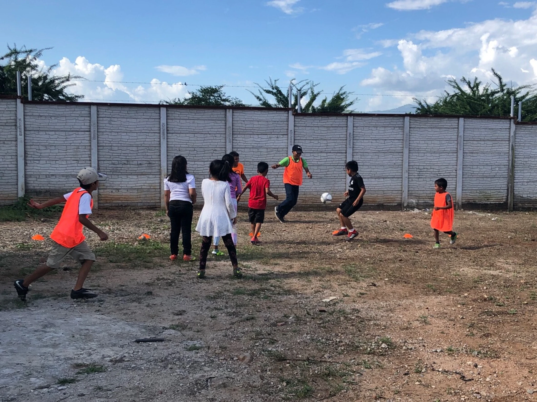 A group of children and an adult play soccer on a dirt field in front of a large concrete wall. Some players wear bright orange vests.