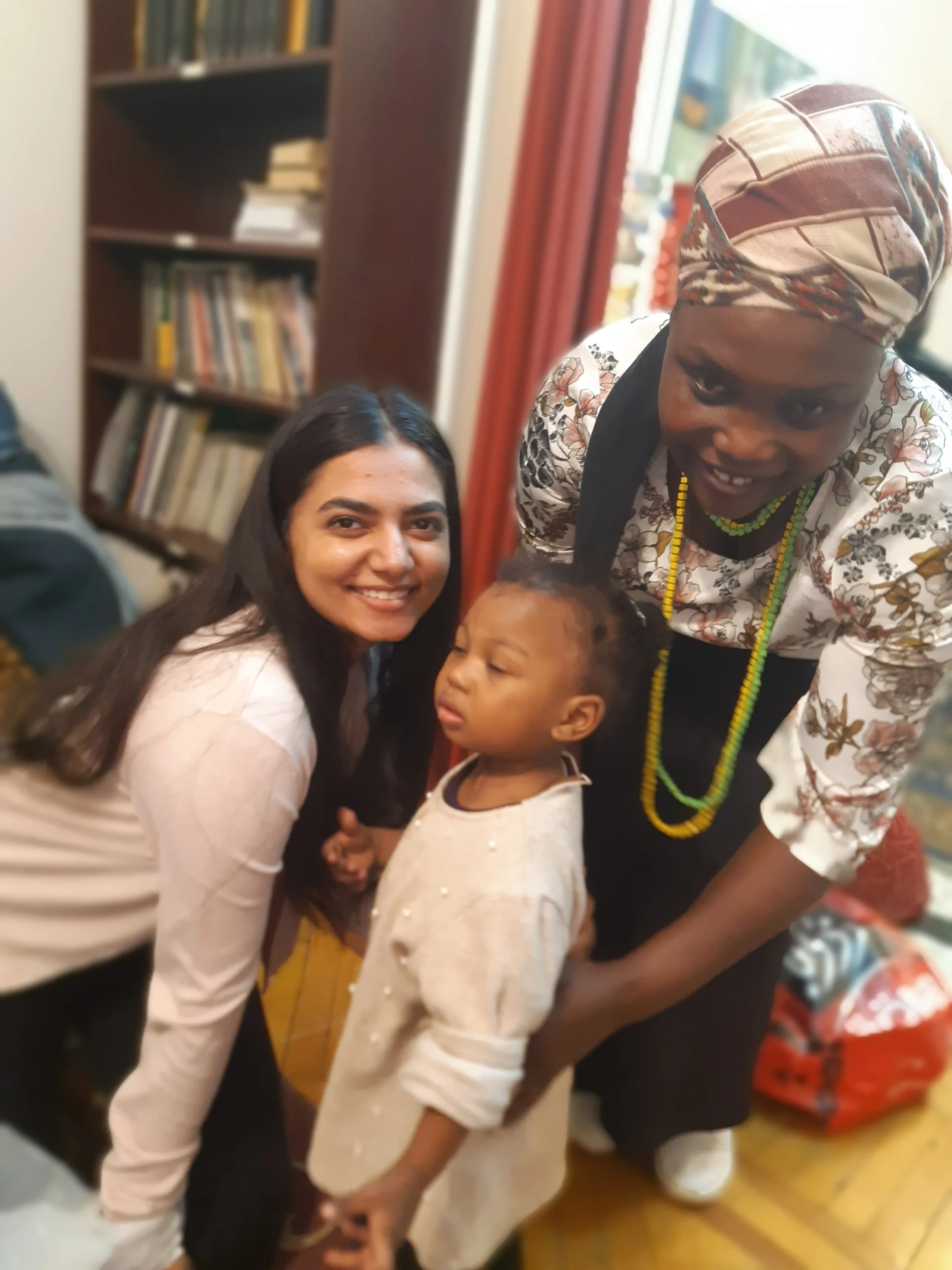 A smiling woman with long dark hair poses with a young child, while another woman in a floral blouse smiles down at them from behind.