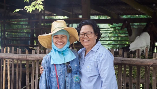 Two smiling women stand together in front of a rustic wooden goat pen. One wears a straw hat and the other wears glasses. Goats are visible behind them.
