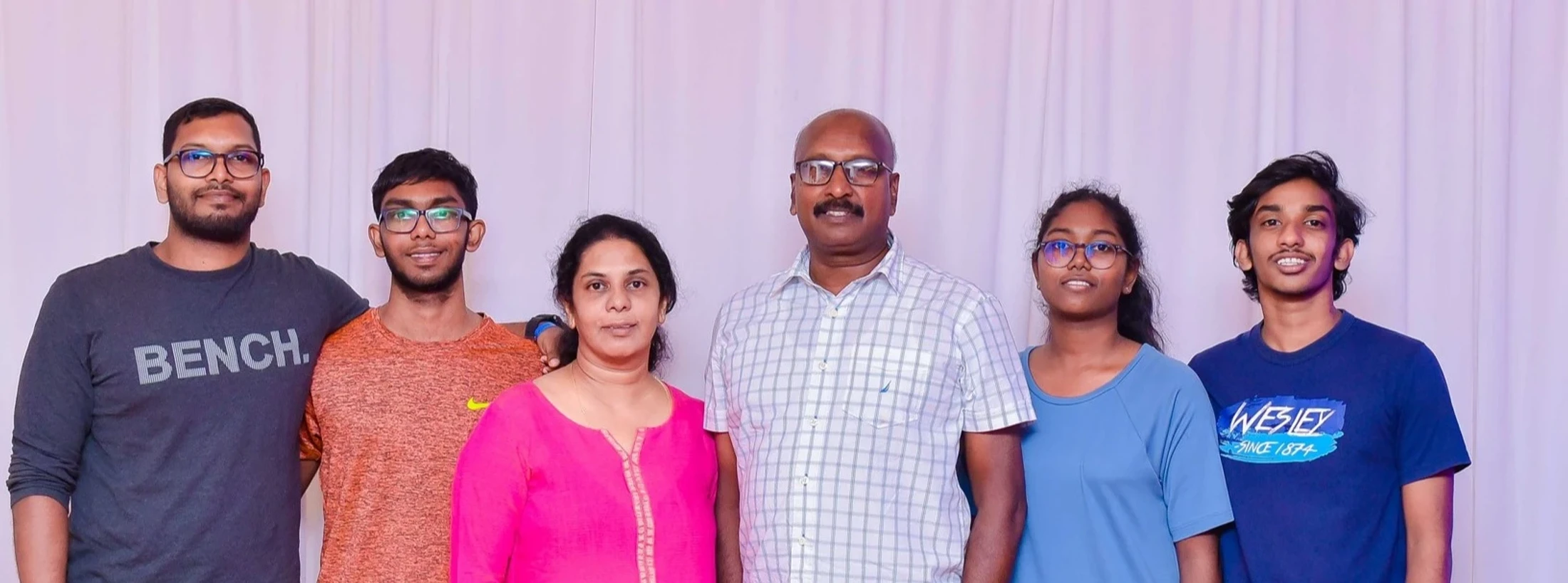 A family portrait of six people, two parents and four young adult children, standing together and smiling in front of a white curtain backdrop.