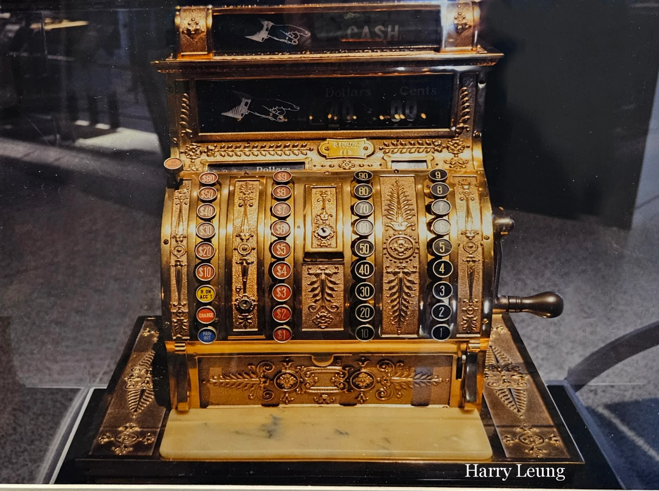 A close-up photograph of an elaborate, vintage brass cash register with multiple columns of numbered keys and intricate, decorative metalwork.