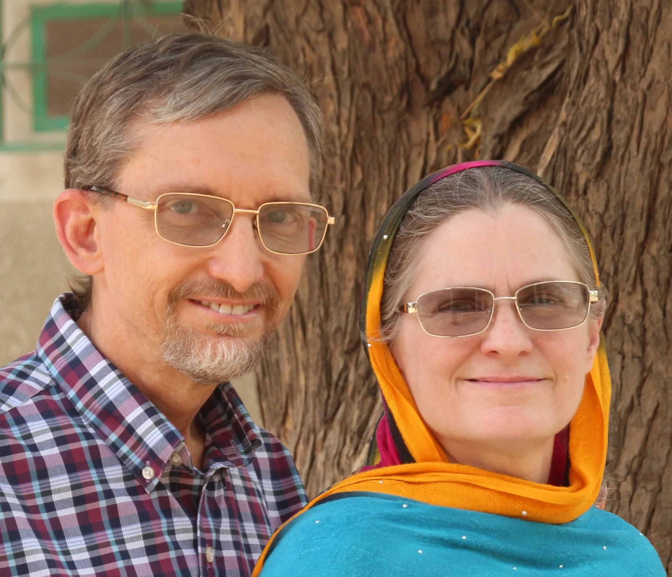 A middle-aged man in a plaid shirt and a woman wearing a colorful headscarf smile for a portrait in front of a large tree.