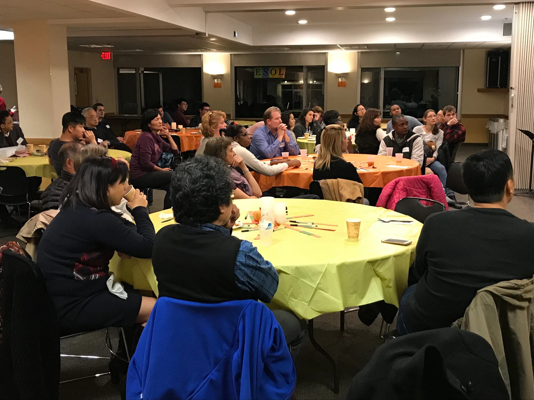 A diverse group of adults sit around yellow and orange covered tables in a large event room, attentively listening to a speaker who is off-camera.