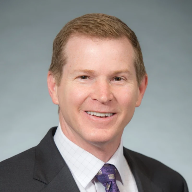 A head and shoulders portrait of a smiling man with light brown hair, wearing a dark suit, patterned shirt, and a purple tie against a gray background.