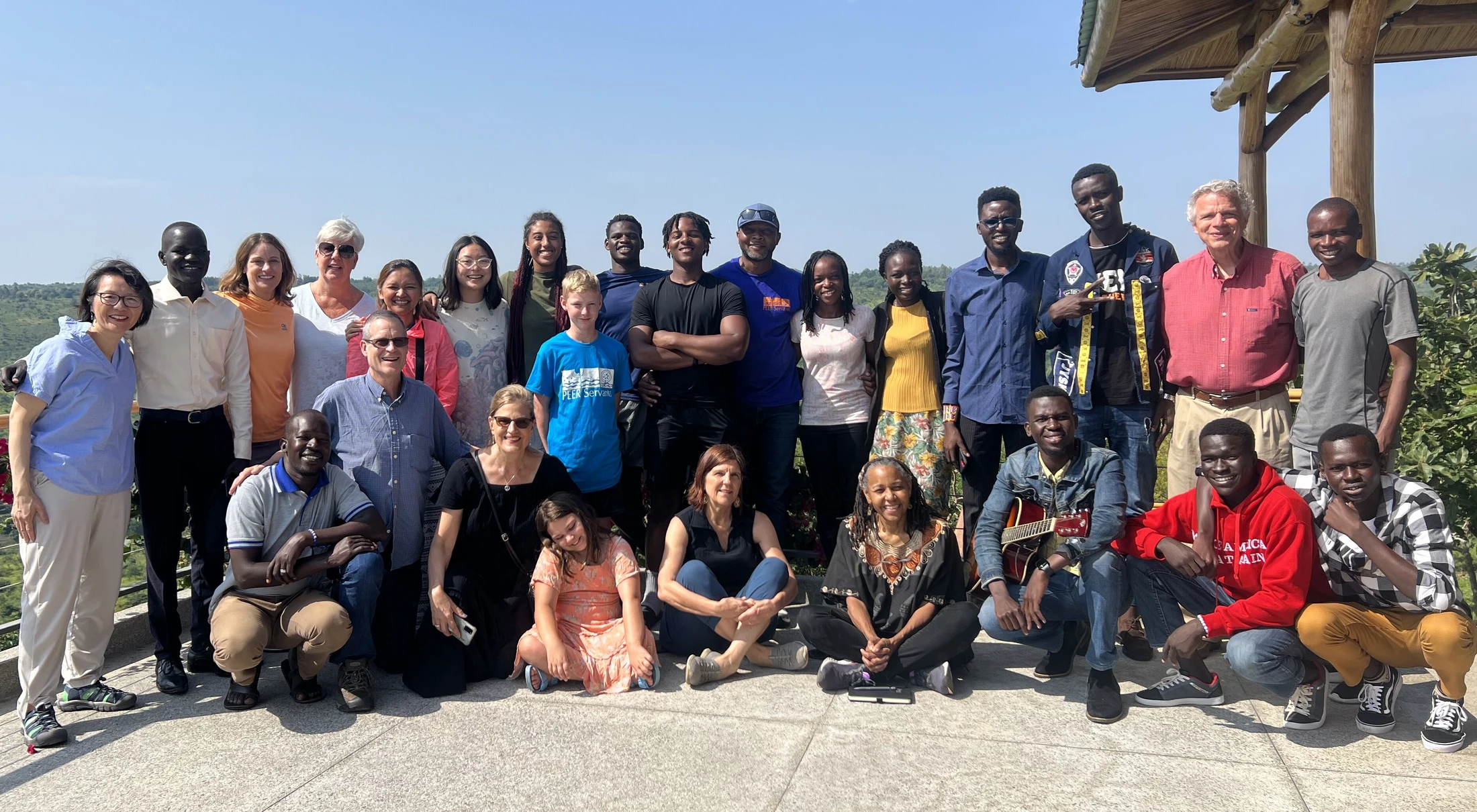A large, diverse group of adults and children smile for a photo on a sunny outdoor patio. A green, hilly landscape is visible in the background.