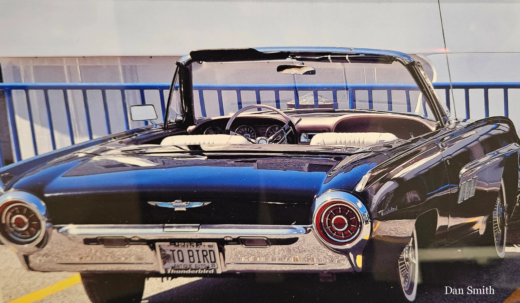 A rear three-quarters view of a shiny black 1963 Ford Thunderbird convertible with a white interior, parked in a lot with a blue railing in the background.