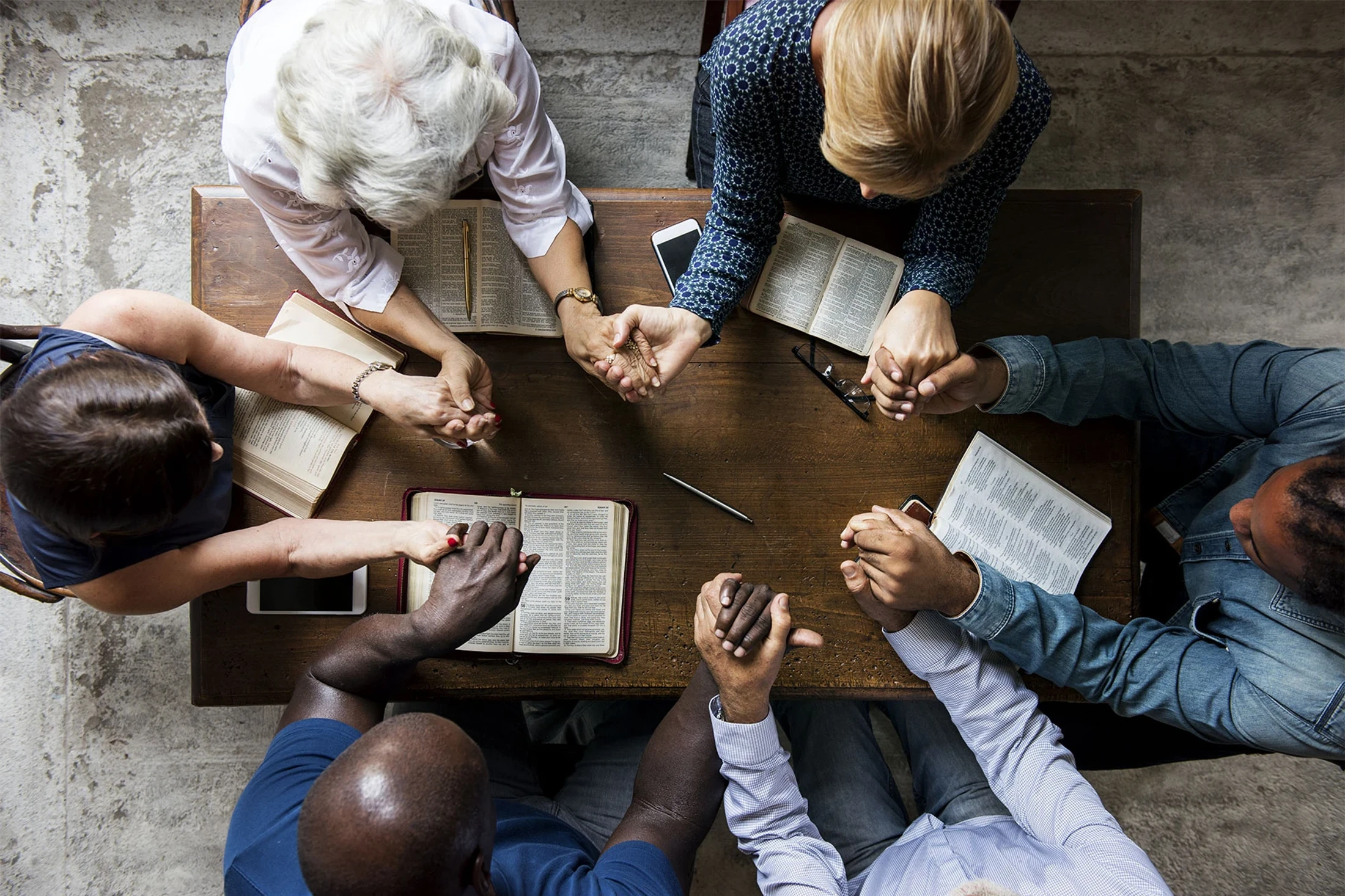 An overhead view of a diverse group of six people sitting around a wooden table, holding hands in prayer with open Bibles in front of them.