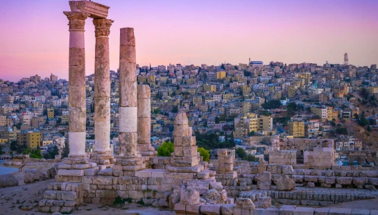 The ancient ruins of the Temple of Hercules at the Amman Citadel, with its tall stone columns, overlook the sprawling city of Amman under a vibrant sunset.