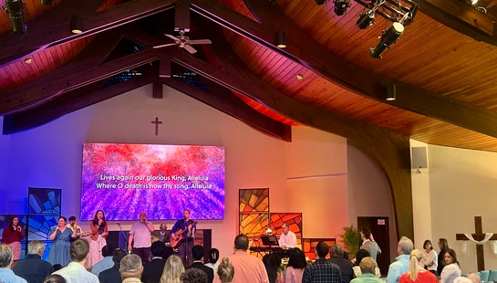 A congregation stands in a church with a high, wood-beamed ceiling, watching a worship band on stage. A large screen displays song lyrics.