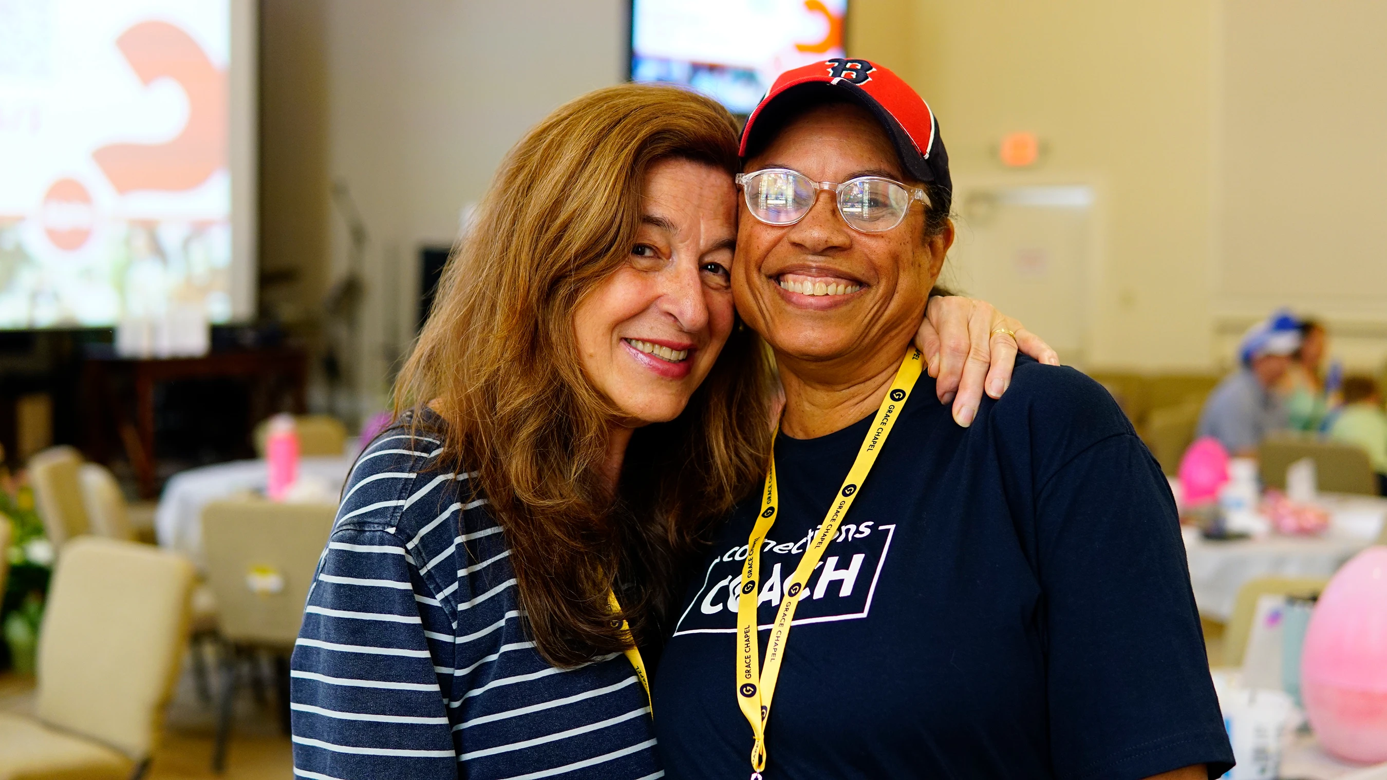 Two women smiling warmly for the camera. One has her arm around the other's shoulder. They are indoors at what looks like a conference or gathering.