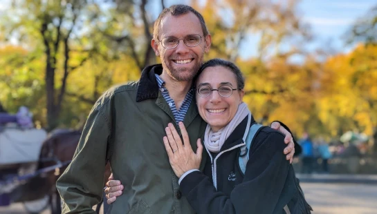 A smiling man and woman wearing glasses embrace while posing for a photo in a park with colorful autumn trees in the background.
