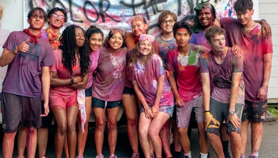 A diverse group of smiling teenagers pose together, their clothes and skin splattered with colorful powder after an event with a 'Color the Night' banner.