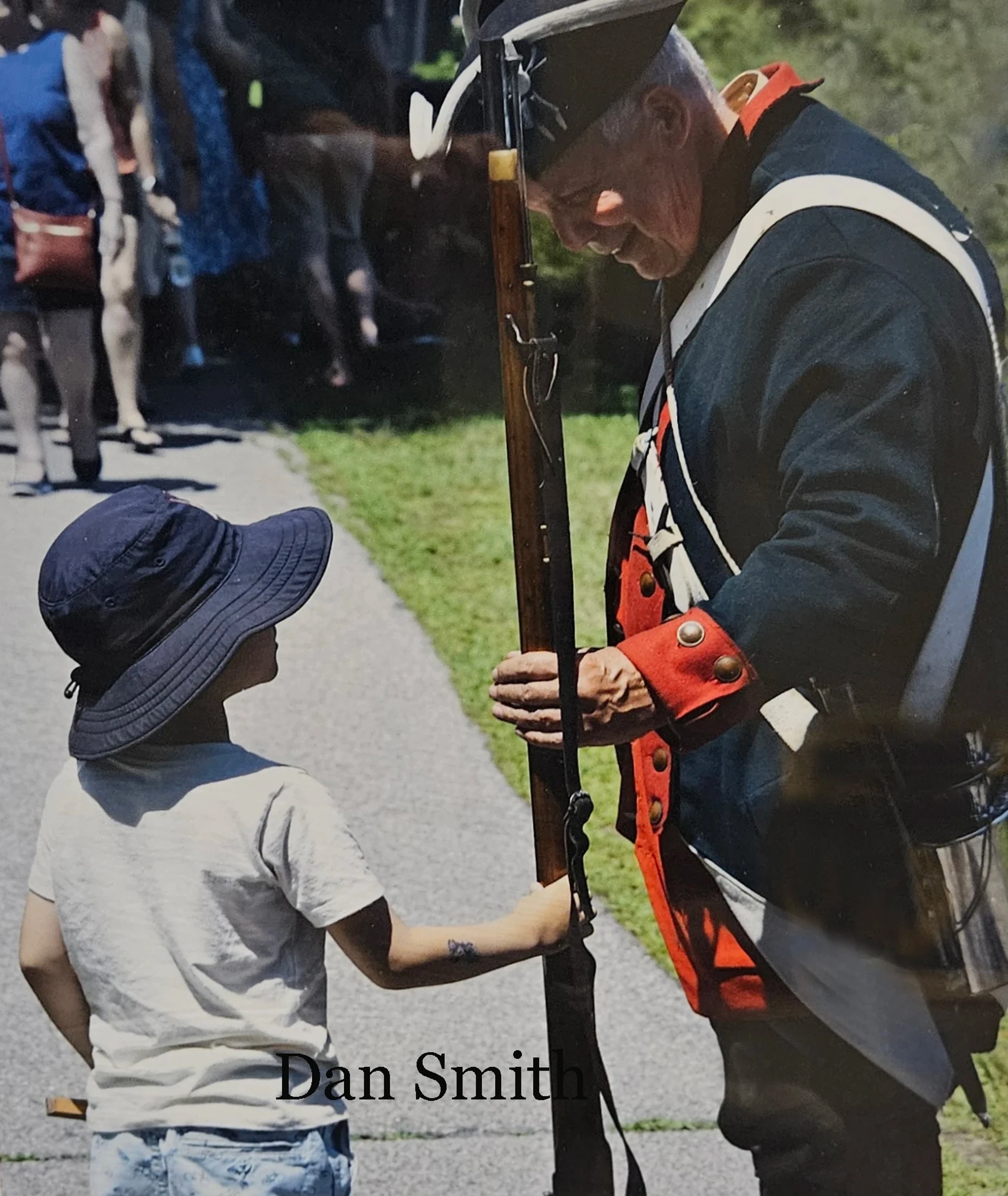 An elderly man in a historical soldier's uniform smiles down at a young boy in a sun hat who is curiously touching his long rifle.