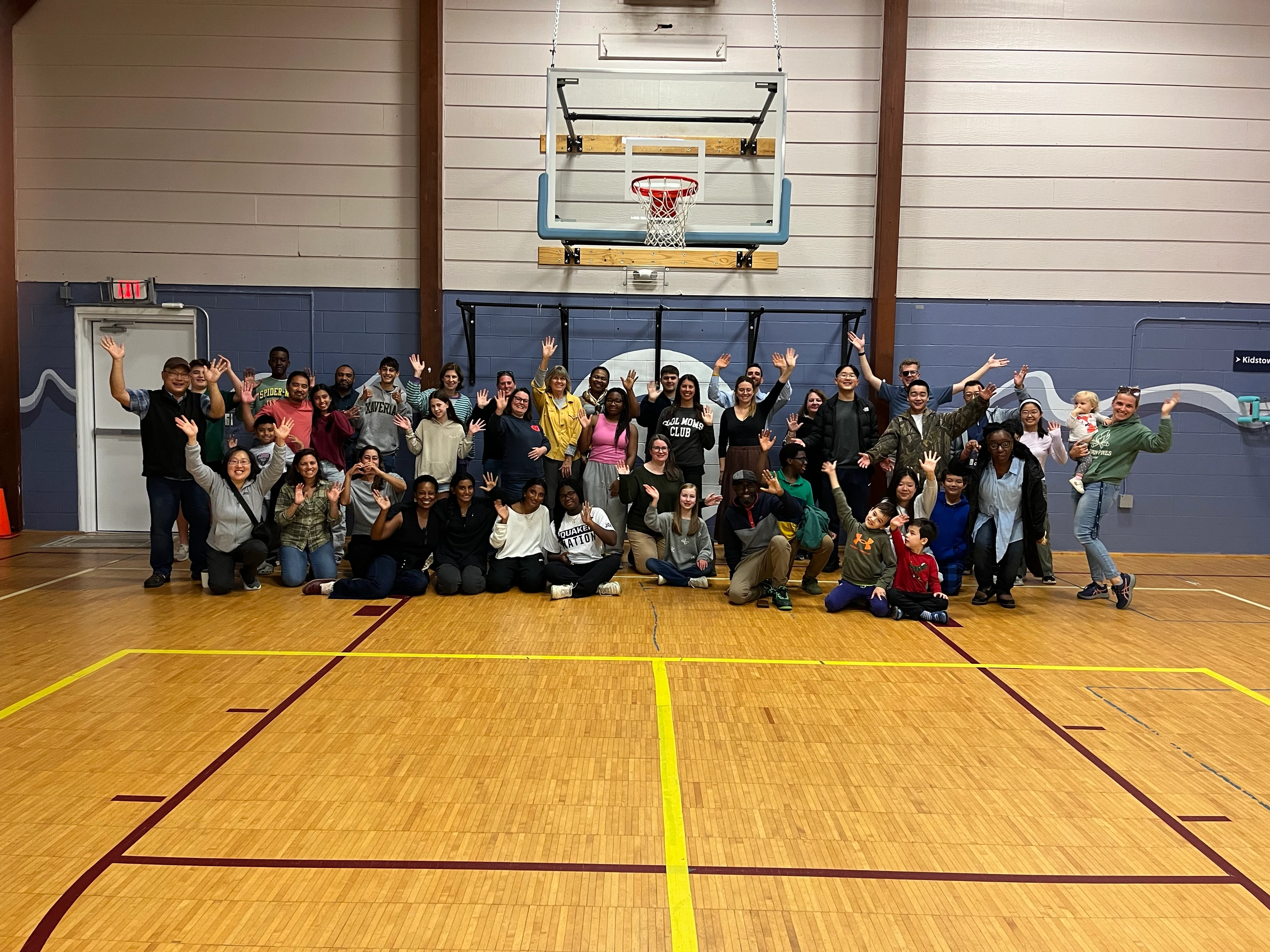 A large, diverse group of smiling adults and children pose together for a photo on an indoor basketball court. Many of them are waving at the camera.