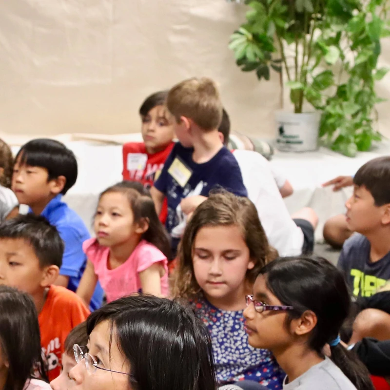A diverse group of elementary school-aged children are sitting on the floor indoors, looking attentively in the same direction at something off-camera.