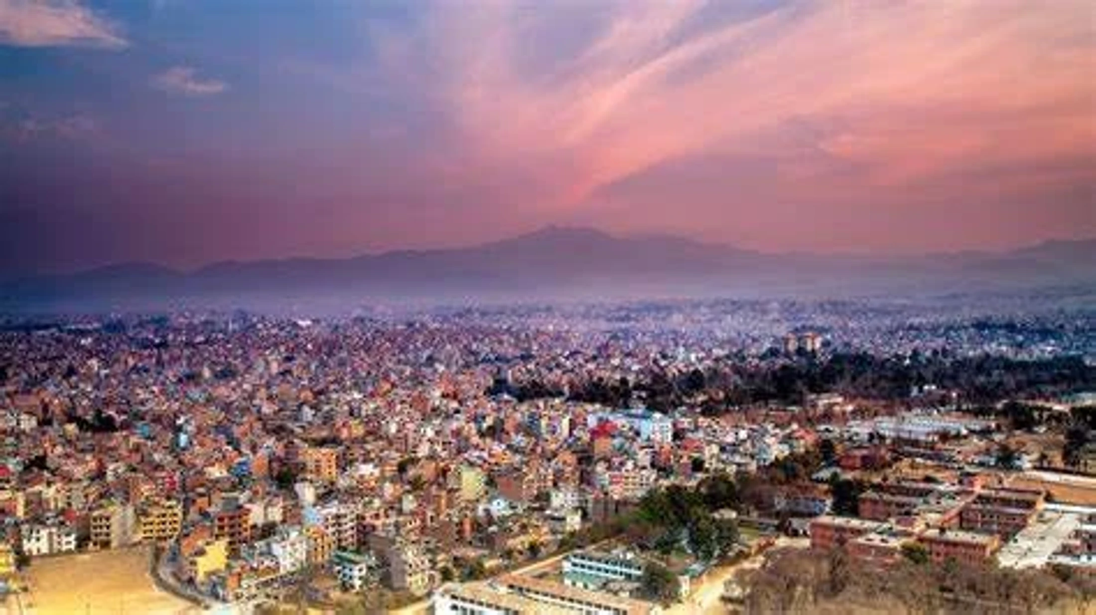 A sprawling, dense cityscape of Kathmandu, Nepal, viewed from above at sunrise, with mountains in the misty background under a pink and purple sky.