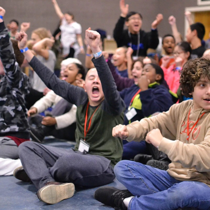 A diverse group of young students sits on a gym floor, cheering excitedly with their fists raised in the air during a school rally or event.